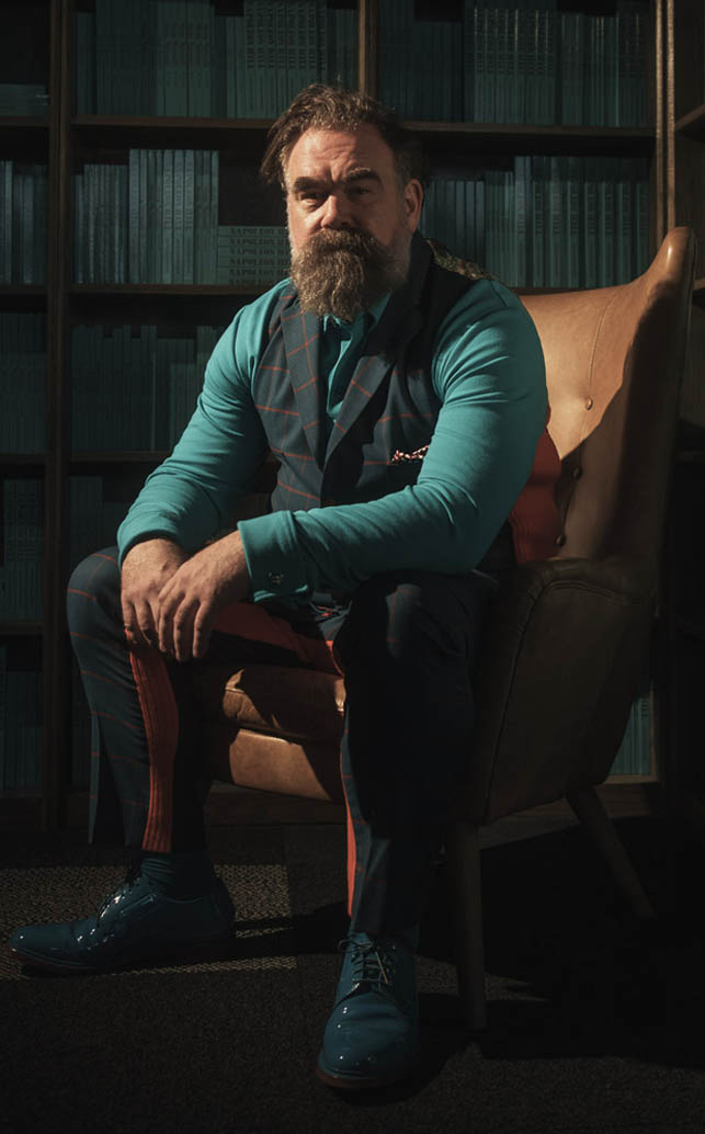 sitting in a library chair wearing a teal outfit under dramatic lighting.