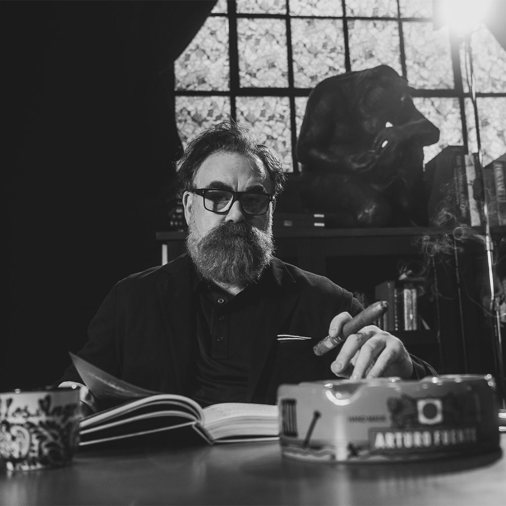 sitting at a desk in a black-and-white office scene, reading a book and holding a cigar with shelves and décor behind him.